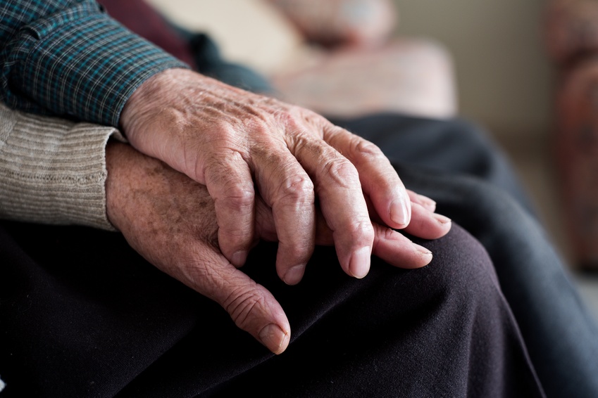 A close-up of two aging adults sitting down together. One has their hand placed on top of the other person's hand.