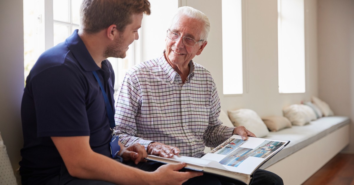 A senior father sits with his adult son as they look at each other and smile. The father holds a photo book.